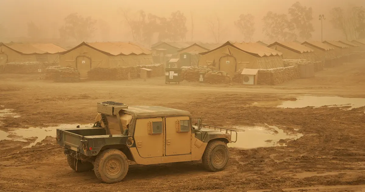 A military truck sits on muddy ground in front of a row of sandbag-reinforced tents during a sandstorm, with dust filling the air.