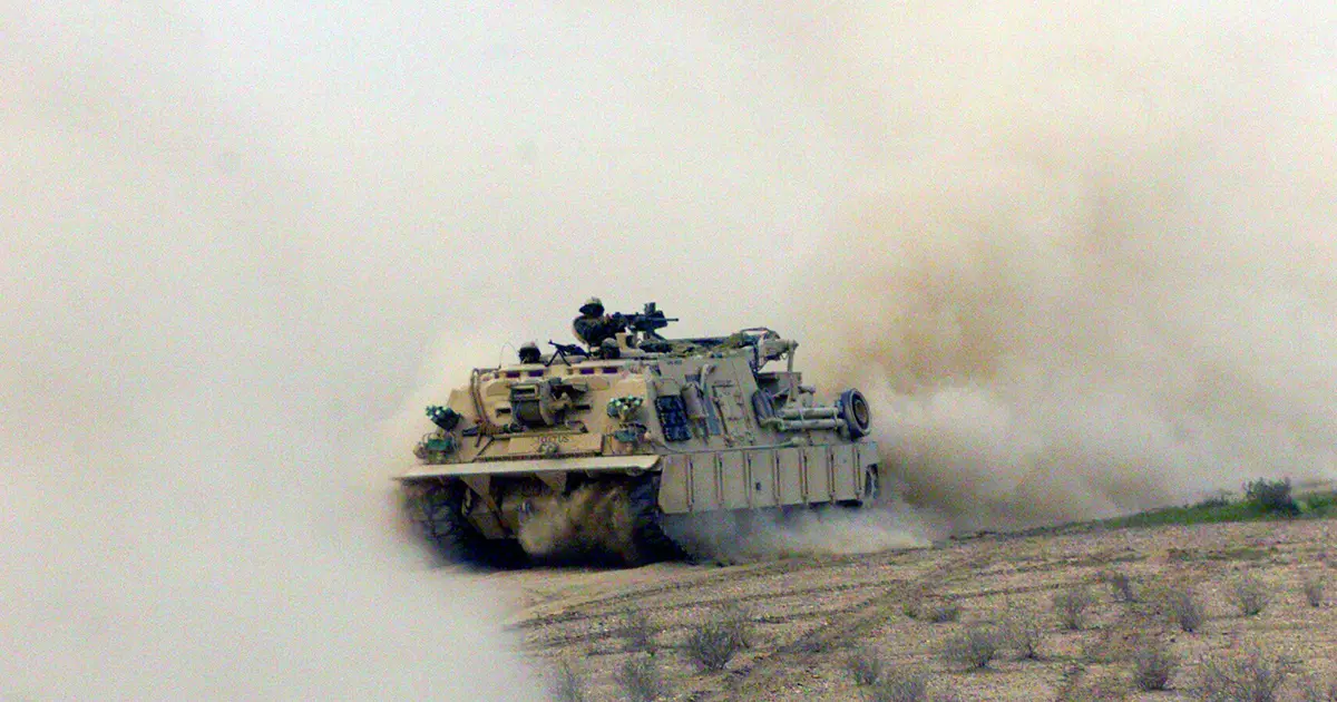 A military armored vehicle moves through a dusty desert terrain, obscured by a large cloud of smoke or dust, with soldiers visible on top, operating a mounted weapon.